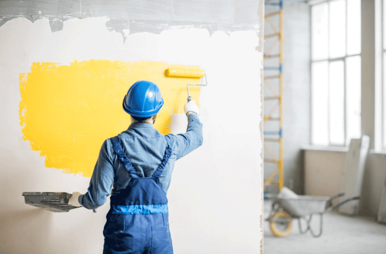 Painter Applying Yellow Paint To A Wall With A Roller, Illustrating The Process Of Painting A House Interior. The Post Is About The Costs Involved In Painting A House Interior.