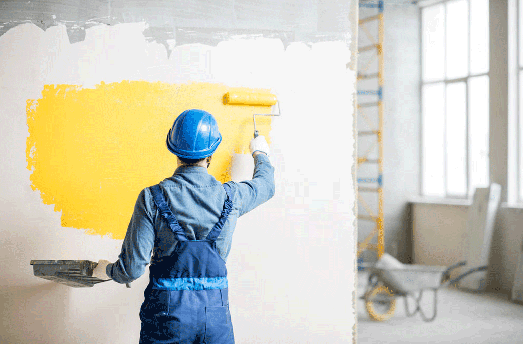 Painter applying yellow paint to a wall with a roller, illustrating the process of painting a house interior. The post is about the costs involved in painting a house interior.